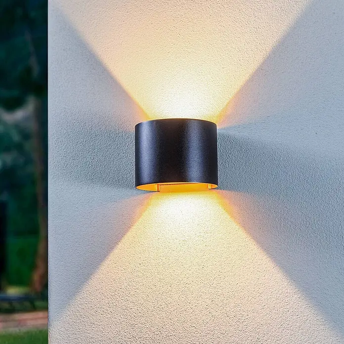 Lindby LED Wandleuchte aussen in schwarz, gold, up down Aussenlampe Wand -  - Schwarze zylindrische Außenwandleuchte, die warmes Licht nach oben und unten auf einer strukturierten Wand abgibt.