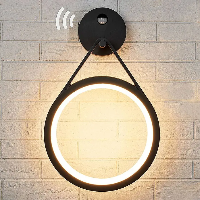 Lucande LED Wandleuchte aussen in graphit, weiß, mit Bewegungsmelder, -  - Eine schwarze runde LED-Wandleuchte mit Bewegungssensor an einer weißen Ziegelwand, die warmweißes Licht abgibt.
