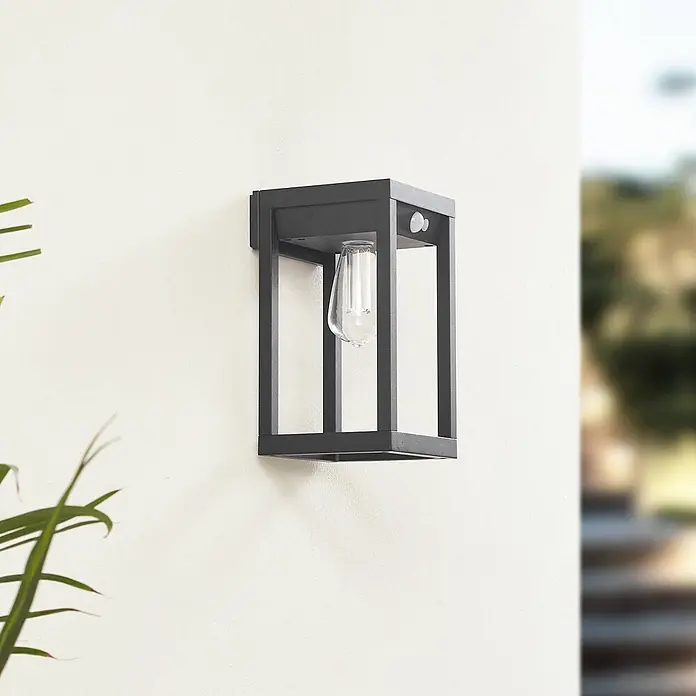 Lindby LED Solar Wandleuchte aussen Smart Home in schwarz, transparent, mit -  - Schwarze moderne Außenwandleuchte mit klarer Glühbirne und Bewegungssensor, montiert an einer hellen Wand.