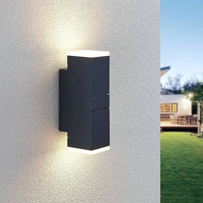 Lindby LED Wandleuchte aussen in anthrazit, weiß satiniert, Aussenlampe -  - Moderne schwarze Außenwandleuchte an einer strukturierten Wand, die nach oben und unten leuchtet, mit Haus und Garten im Hintergrund.