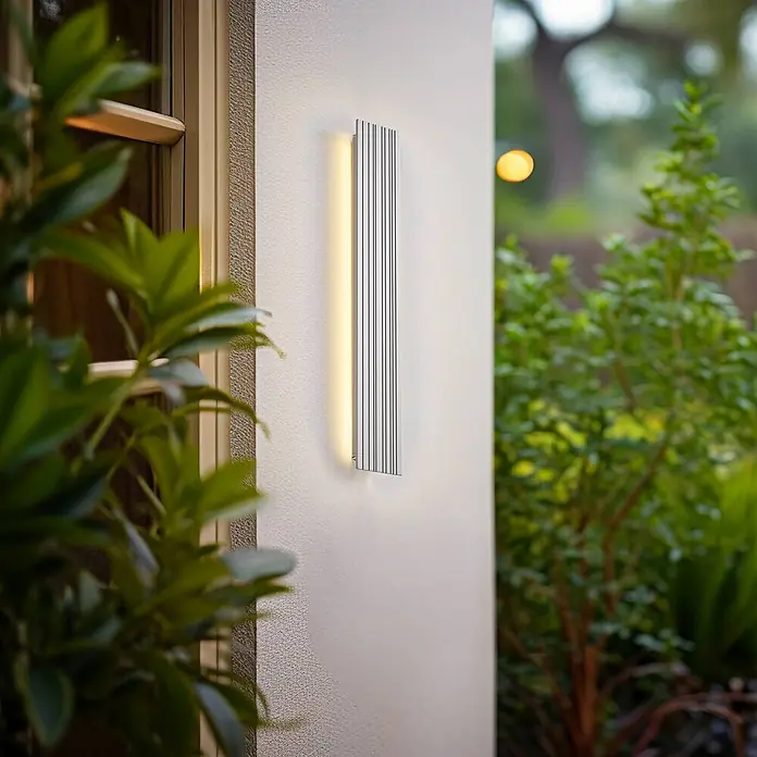 Lucande Außenwandleuchte Melira - alu / grau / zink, alu / grau / zink, IP65 - Silberne rechteckige Außenwandleuchte mit vertikalen Rillen, die warmes Licht auf eine Wand im Garten strahlt.