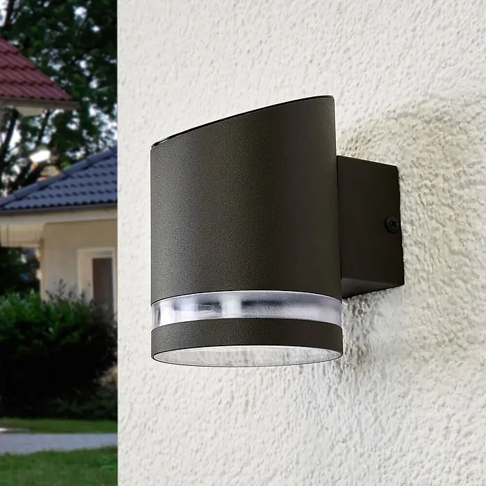 Lindby Solar LED Wandlampe / Wandleuchte außen, 1x 0,5W LED, Downlight -  - Dunkelgraue zylindrische Außenwandleuchte mit transluzentem Band, an einer strukturierten Außenwand montiert.