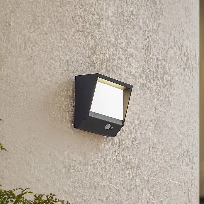 Lucande Außenwandleuchte Dava - schwarz, IP54 - Anthrazitfarbene Außenwandleuchte mit Bewegungssensor, strahlt warmweißes Licht an einer strukturierten Wand aus.