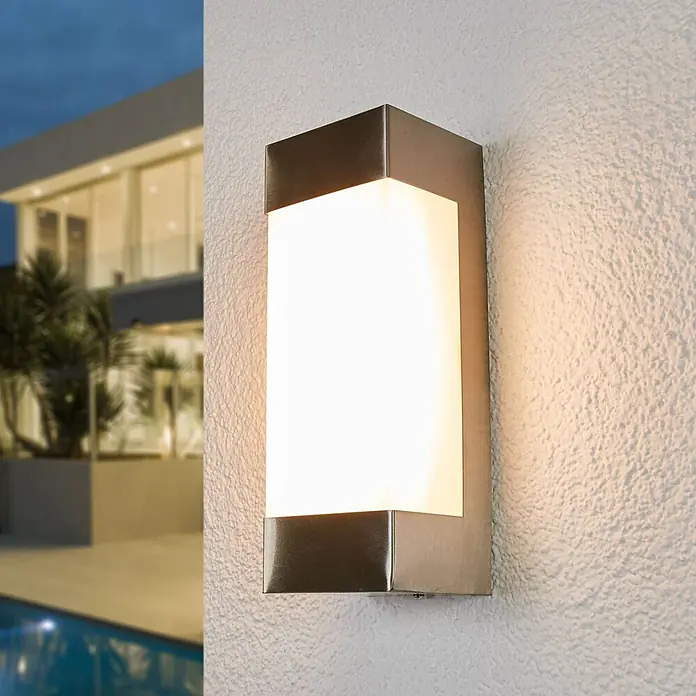 Lindby Edelstahl Wandlampe aussen, 1x 6,5W LED, Außenleuchte -  - Beleuchtete rechteckige Außenwandleuchte aus gebürstetem Edelstahl mit milchweißem Diffusor, beleuchtet eine strukturierte Wand vor einem modernen Haus.