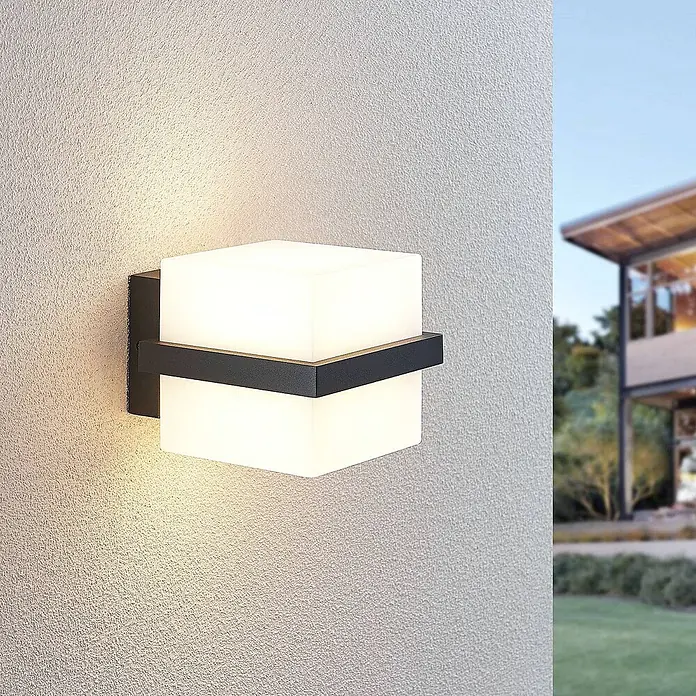 Lindby LED Wandleuchte aussen in anthrazit, weiß, Aussenlampe Wand -  - Moderne schwarze Außenwandleuchte mit kubischem weißem Diffusor, die ein warmes Licht auf eine strukturierte Wand wirft.