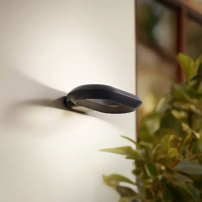 Lucande Außenwandleuchte Loop - schwarz, IP54 - Moderne schwarze Wandleuchte mit organischem Design, an einer hellen Wand montiert, für indirekte Beleuchtung.