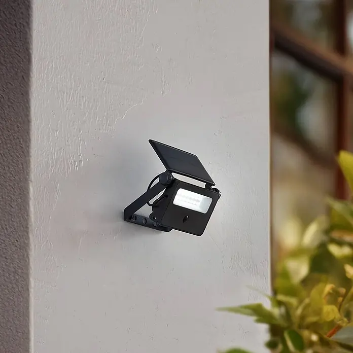 Lindby LED Solar Wandleuchte aussen in schwarz, mit Bewegungsmelder, -  - Schwarzer Solar-LED-Flutstrahler an Außenwand montiert, Solarpanel nach oben und Lichtfeld sichtbar.