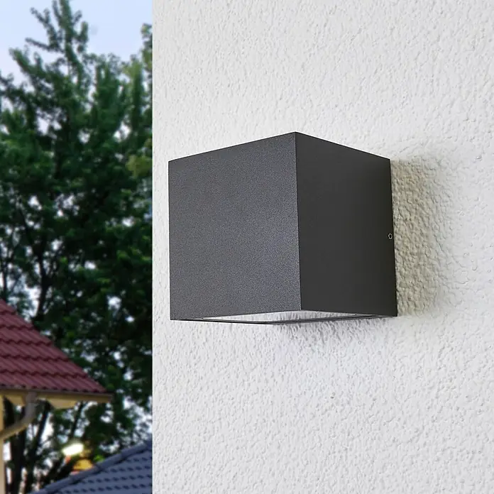 Lucande LED Wandleuchte aussen in dunkelgrau, Aussenlampe Wand -  - Dunkelgraue, würfelförmige Außenwandleuchte an einer strukturierten weißen Wand, mit Baum und Dach im Hintergrund.