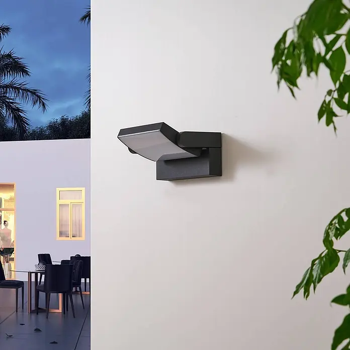 Lindby LED Wandleuchte aussen 340° schwenkbar, Wandlampe aussen -  - Schwarze Außenwandleuchte in einer Abendszene mit Palmen und einer beleuchteten Terrasse.