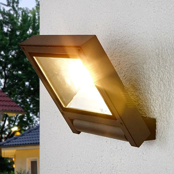 Lucande LED Wandleuchte innen in anthrazit, Wandstrahler -  - Braune, angewinkelte Außenwandleuchte an einer strukturierten Wand, die helles, warmes Licht abstrahlt. Zeigt ein Haus im Freien in der Dämmerung.