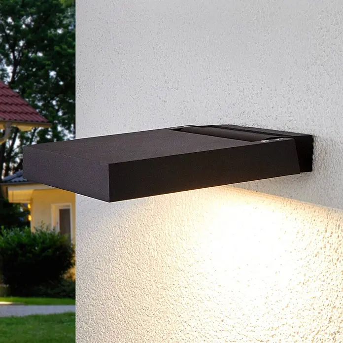 Lucande LED Wandleuchte innen in anthrazit, Wandstrahler -  - Eine schwarze, rechteckige Außenwandleuchte an einer strukturierten Wand, die ein warmes Licht nach unten wirft.