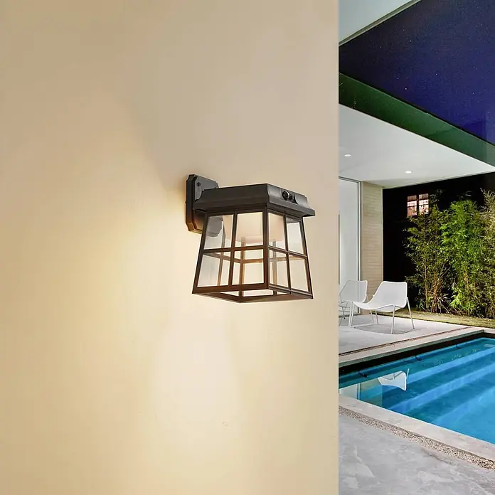 Lindby LED Solar Wandleuchte aussen in schwarz, mit Bewegungsmelder, -  - Klassische dunkle Laternen-Wandleuchte für den Außenbereich mit Glasscheiben, beleuchtet und an einer hellen Wand neben einem Pool montiert.