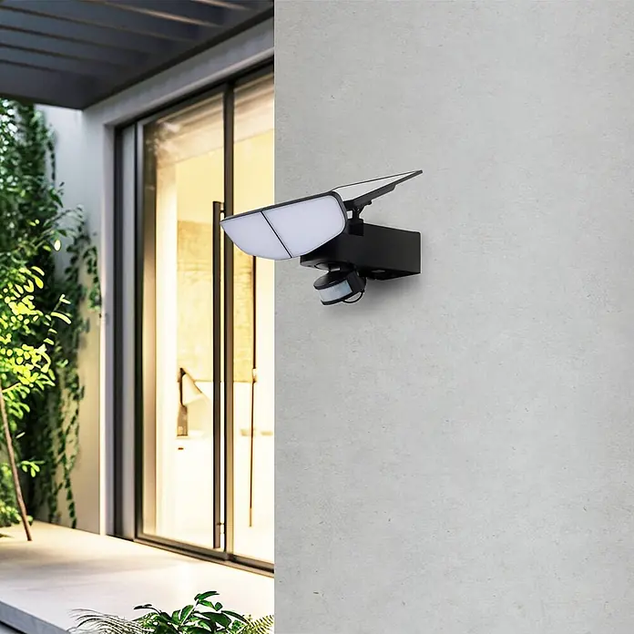 Lindby Außenwandleuchte Naelin - schwarz, schwarz, IP44 - Schwarze Solar-Außenleuchte mit verstellbaren LED-Paneelen und Bewegungssensor an einer hellgrauen Wand neben einer Glastür.