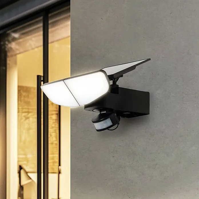 Lindby Außenwandleuchte Naelin - schwarz, schwarz, IP44 - Schwarzer solarbetriebener LED-Fluter mit Bewegungssensor, an grauer Betonwand neben Glastür montiert.