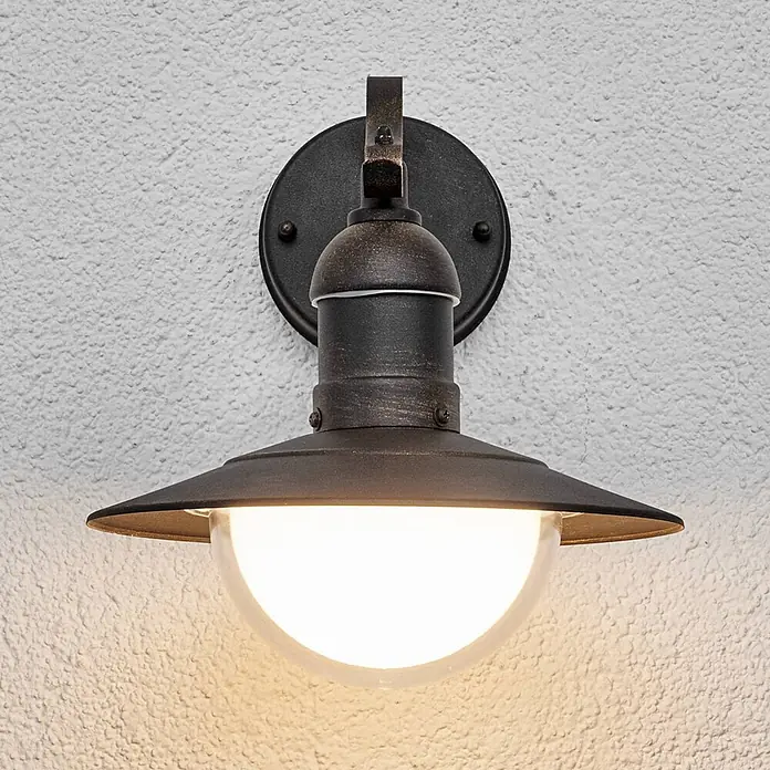 Lindby Edelstahl LED Wandlampe aussen Antik Look, LED Wandleuchte außen -  - Wandleuchte aus gealterter Bronze mit klarem Glasdiffusor, beleuchtet und an einer strukturierten Wand montiert.
