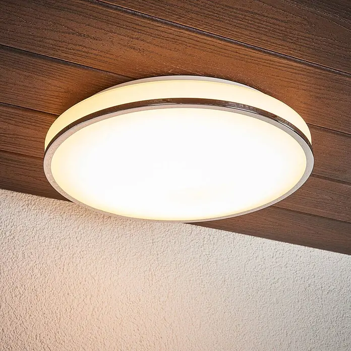 Lindby LED Deckenlampe in weiß, chrom, Deckenleuchte rund -  - Runde LED-Deckenleuchte mit silbernem Rand und zusätzlichem Leuchtring, an einer Holzdecke über einer strukturierten Wand montiert.