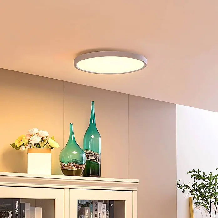Lindby LED Deckenlampe Smart Home in weiß, dimmbar Deckenleuchte rund -  - Eine runde LED-Deckenleuchte mit weißem Rahmen, in einem Wohnzimmer installiert, das dekorative Vasen und Möbel warm beleuchtet.