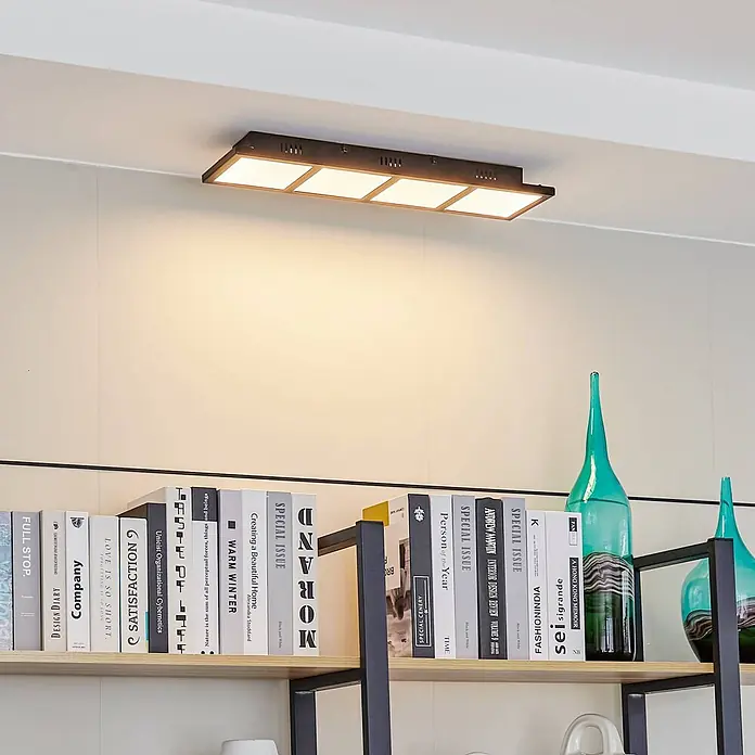 Lindby Deckenleuchte Raymie - schwarz - Rechteckige Deckenleuchte mit dunklem Rahmen und vier quadratischen Leuchtpaneelen, an der Decke über einem Bücherregal montiert.