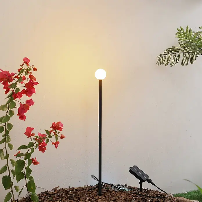 Lindby LED Solar Gartenlampe Erdspieß in schwarz matt, Erdspießlampe -  - Schwarze Solar-Gartenleuchte mit Kugel-Lampe, beleuchtet ein Blumenbeet mit Mulch und Blumen.