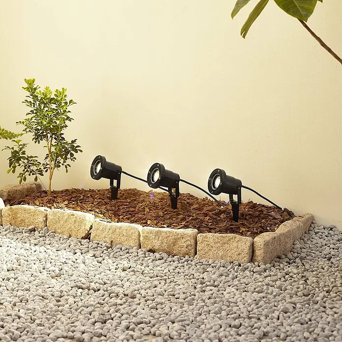 Lindby Gartenstrahler mit Erdspieß in schwarz, Strahler Garten -  - Drei schwarze Gartenspots beleuchten ein gemulchtes Beet mit kleiner Pflanze, das an einen hellen Kiesweg vor einer cremefarbenen Wand grenzt.