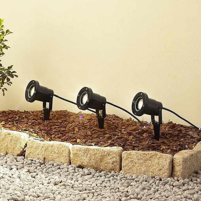 Lindby Gartenstrahler mit Erdspieß in schwarz, Strahler Garten -  - Drei schwarze Gartenstrahler beleuchten eine Wand in einem Mulchbeet.