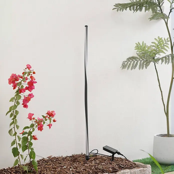 Lindby Erdspießleuchte Sabiel -  - Hohe, schlanke Solar-Gartenleuchte mit gedrehtem Design, installiert in Holzspänen neben rosa Blumen.