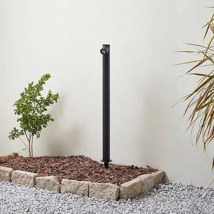Lindby LED Gartenstrahler mit Erdspieß in anthrazit, Strahler Garten -  - Schwarzer Außenstrahler in einem Gartenbeet mit Mulch, umrandet von Steinen, neben einer Pflanze.