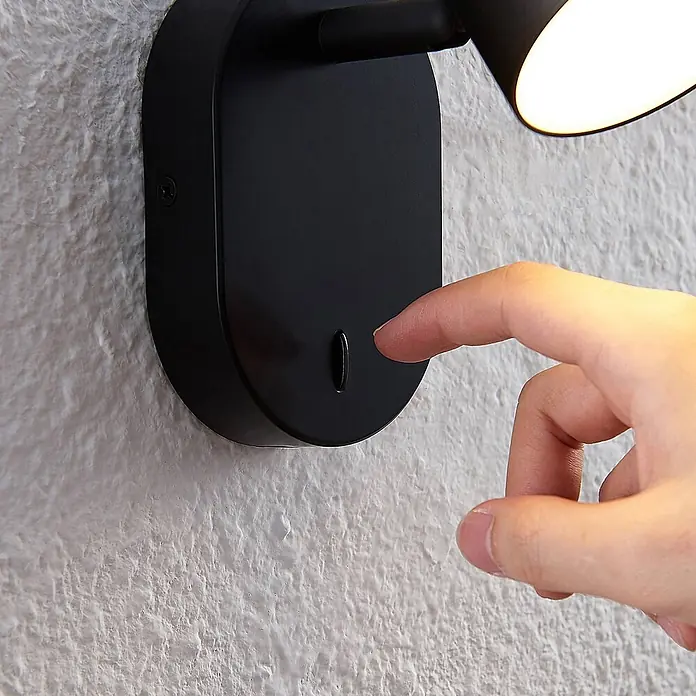 Lindby LED Deckenlampe in schwarz, Deckenstrahler -  - Schwarze Wandleuchte mit eingeschaltetem Licht, ein Finger bedient den Knopfschalter.