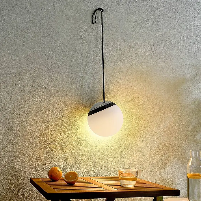 Lindby LED Hängelampe außen in weiß, schwarz, dimmbar Pendelleuchte -  - Runde Hängelampe mit weißem Kugeldiffusor und schwarzem Band, die eine strukturierte Wand über einem Holztisch beleuchtet.