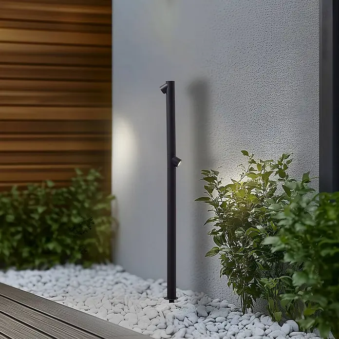 Lindby Erdspießleuchte Luzian -  - Schwarze Outdoor-Pollerleuchte mit zwei verstellbaren Strahlern, beleuchtet strukturierte Wand und Gartenpflanzen.