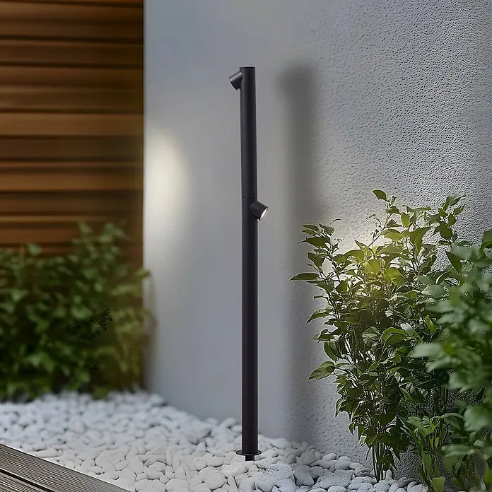 Lindby Erdspießleuchte Luzian -  - Schlanke, schwarze Außenpollerleuchte mit zwei verstellbaren Strahlern, die eine strukturierte Wand und umliegende Pflanzen auf weißen Kieselsteinen beleuchtet.