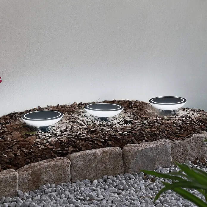 Lindby LED Gartenlampe Erdspieß Smart Home in schwarz, weiß, dimmbar -  - Drei runde, weiß-schwarze Außenleuchten mit leuchtenden Rändern sind in Rindenmulch installiert und beleuchten einen Gartenbereich.