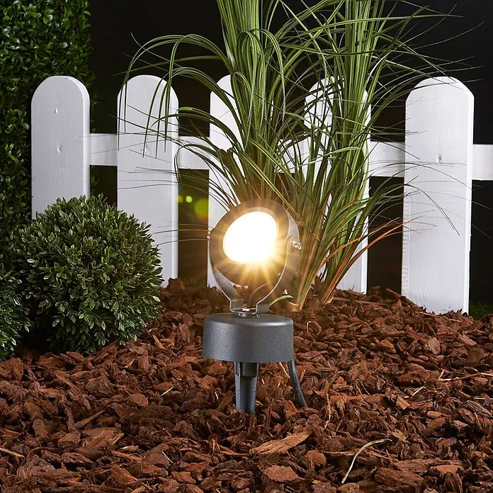 Lucande LED Gartenstrahler mit Erdspieß in anthrazit, Strahler Garten -  - Dunkelgrauer Outdoor-Strahler mit hellem Licht, in einem Gartenbeet mit Rindenmulch installiert, beleuchtet eine Pflanze.