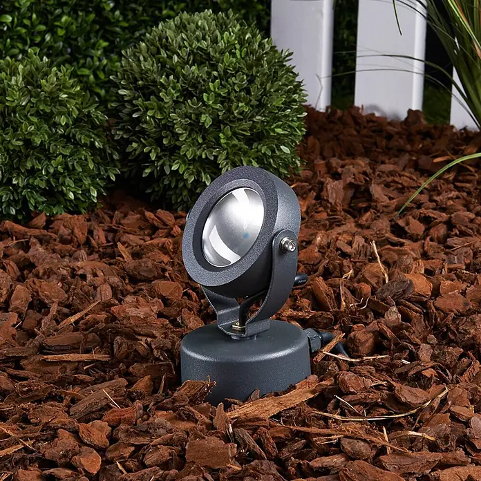 Lucande LED Gartenstrahler mit Erdspieß in anthrazit, Strahler Garten -  - Dunkelgrauer Gartenstrahler auf Rindenmulch, mit grünen Büschen und weißem Zaun im Hintergrund.
