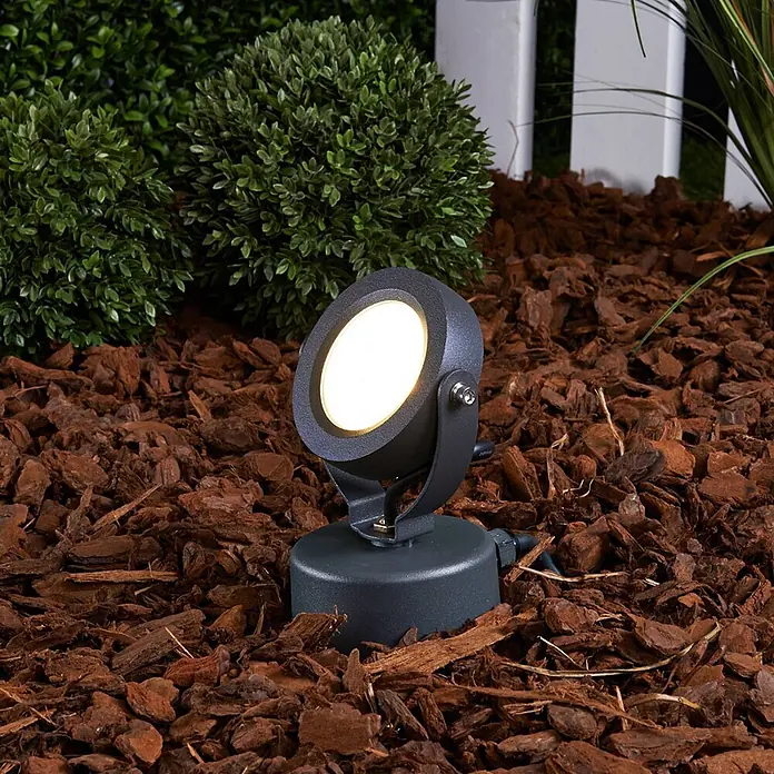Lucande LED Gartenstrahler mit Erdspieß in anthrazit, Strahler Garten -  - Dunkelgrauer Außenstrahler mit warmem Licht, in braunem Rindenmulch umgeben von grünem Laubwerk.