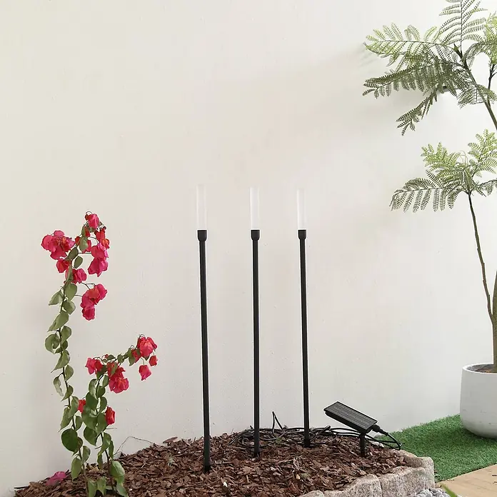 Lindby Erdspießleuchte Balian -  - Drei schwarze Solar-Wegeleuchten mit weißen Diffusoren, in einem Gartenbeet mit separatem Solarpanel installiert.