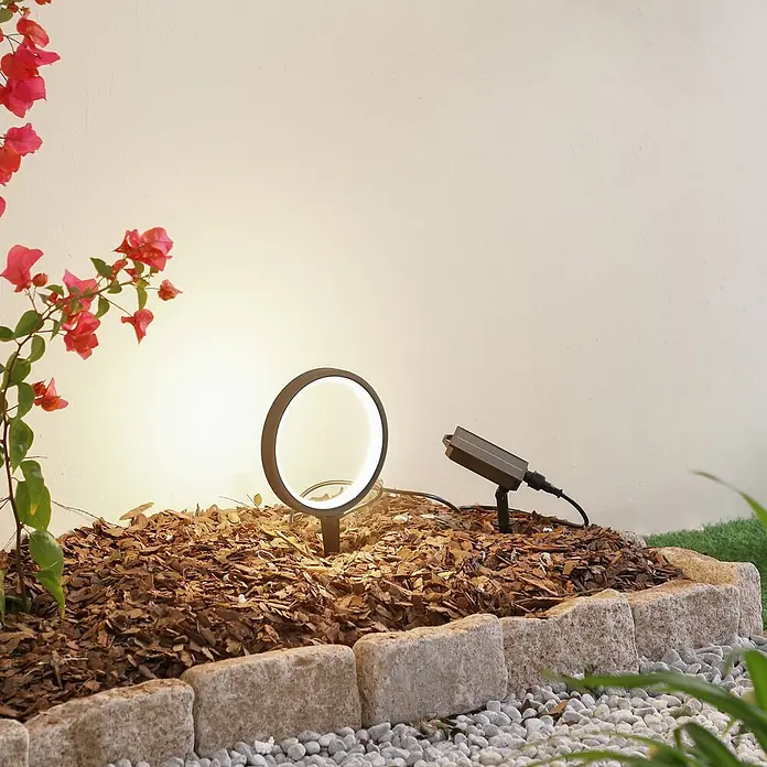 Lindby Erdspießleuchte Syrene -  - Schwarzer solarbetriebener Gartenstrahler und Solarpanel, beleuchten ein Gartenbeet mit Rindenmulch und Blumen.