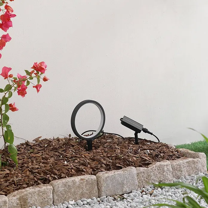 Lindby LED Solar Gartenlampe Erdspieß in schwarz matt, Erdspießlampe -  - Schwarze runde LED-Solar-Gartenleuchte mit angeschlossenem Solarpanel, in einem Mulchbeet.