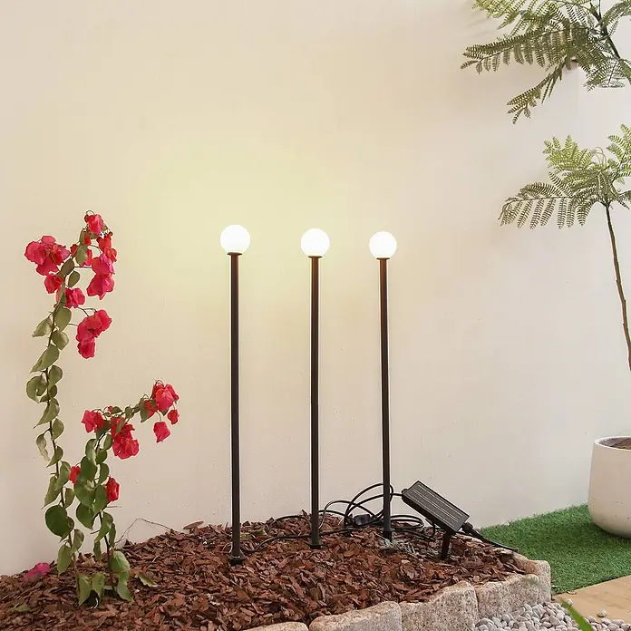 Lindby LED Solar Gartenlampe Erdspieß in schwarz matt, Erdspießlampe -  - Drei solarbetriebene Gartenleuchten mit schwarzen Stielen und weißen Kugeln, in einem Mulchbeet neben blühenden Pflanzen.
