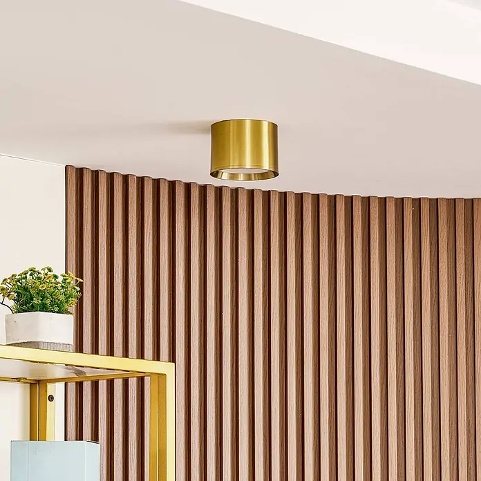 Lindby LED Aufbauleuchte gold Ø 12 cm, Aufbaustrahler, Deckenspot 15W LED -  - Eine zylindrische Deckenleuchte aus Messing ist an einer weißen Decke montiert und beleuchtet einen modernen Raum mit einer Lattenholzwand.