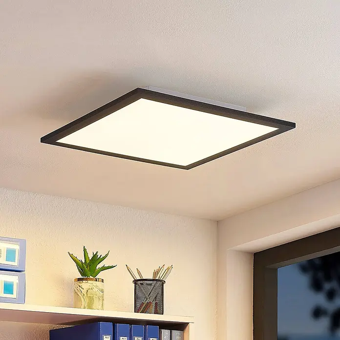 Lindby LED-Panel Nelios - schwarz, schwarz, Neutralweiß - Eine quadratische LED-Deckenleuchte mit dunklem Rahmen beleuchtet einen modernen Raum mit Regal und Fenster.