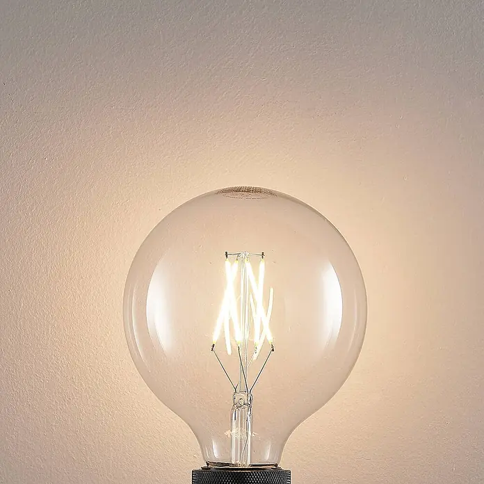 Arcchio Leuchtmittel -  - Große, klare LED-Globusbirne mit sichtbaren Filamenten, die warmes Licht auf eine strukturierte Wand wirft.