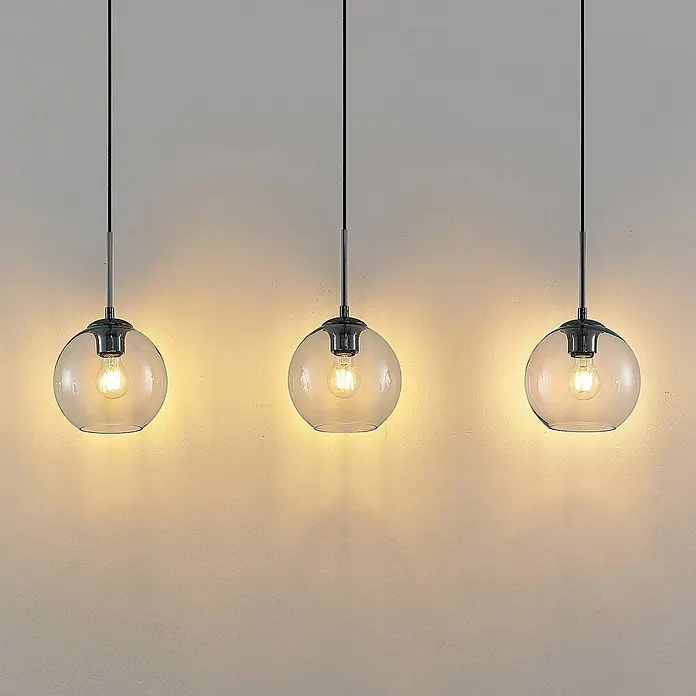 Lindby Pendelleuchte in transparent, Hängelampe -  - Drei Pendelleuchten mit klaren Glaskugeln und dunklen Metallfassungen, beleuchtet und warmes Licht an eine Wand werfend.