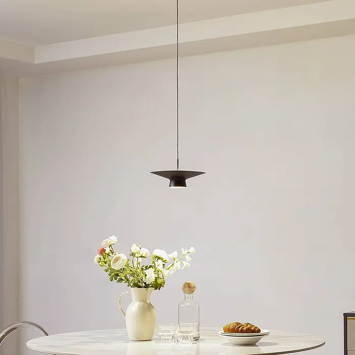 Lindby Pendelleuchte Norvind - schwarz, sandSchwarz, gold - Eine moderne schwarze kegelförmige Pendelleuchte beleuchtet einen Esstisch mit Blumen und Frühstück.