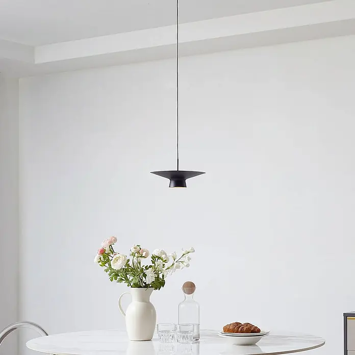 Lindby Pendelleuchte Norvind - schwarz, sandSchwarz, gold - Eine schwarze, scheibenförmige Pendelleuchte hängt elegant über einem weißen Esstisch mit Blumen und Erfrischungen.