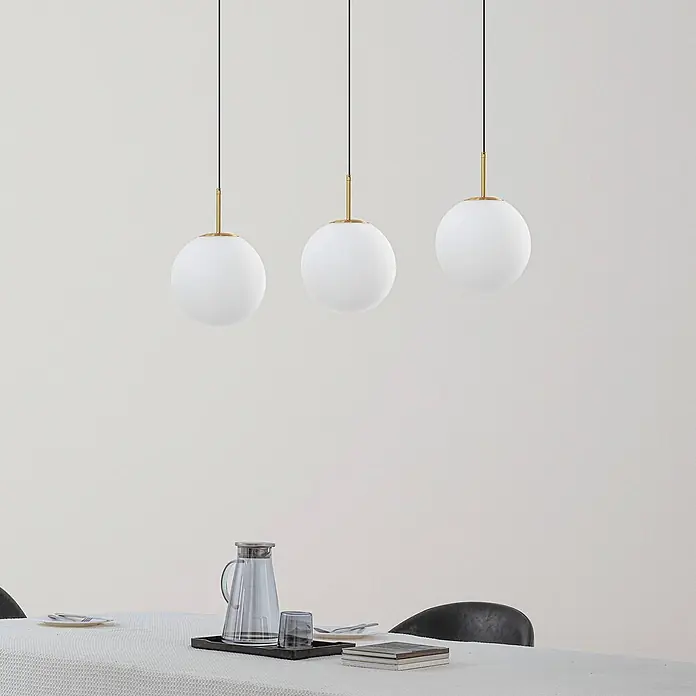 Lucande LED Pendelleuchte in weiß, gold, Hängelampe -  - Drei Pendelleuchten mit weißen Kugelschirmchen und Messingakzenten hängen über einem Esstisch.