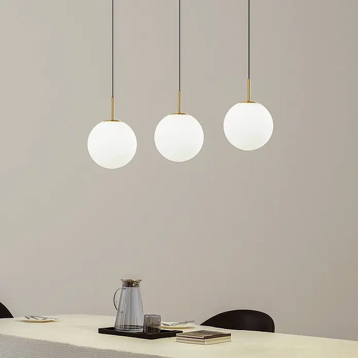 Lucande LED Pendelleuchte in weiß, gold, Hängelampe -  - Drei moderne kugelförmige Pendelleuchten mit weißen Glasschirmen und Messingdetails hängen über einem Esstisch.