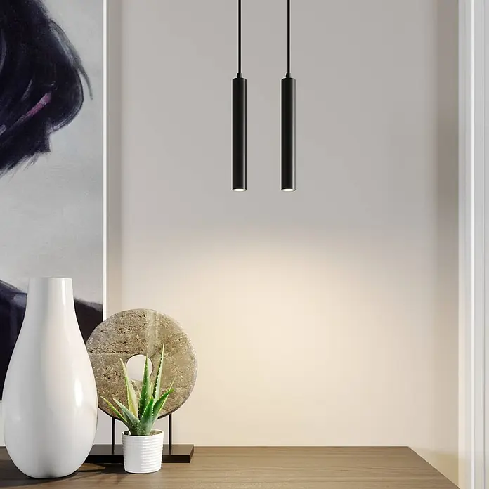 Arcchio LED Pendelleuchte in schwarz, Hängelampe -  - Zwei schlanke, schwarze zylindrische Pendelleuchten hängen von der Decke und werfen einen warmen Schein auf eine Holzoberfläche mit Dekoration.