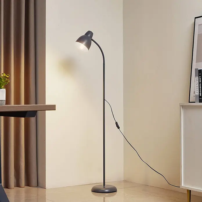 Lindby Stehlampe Wohnzimmer in grau, Stehleuchte -  - Eine dunkelgraue Stehlampe mit verstellbarem Kopf steht in einem Raum und spendet warmes Licht. Sie hat ein sichtbares Stromkabel mit Schalter.