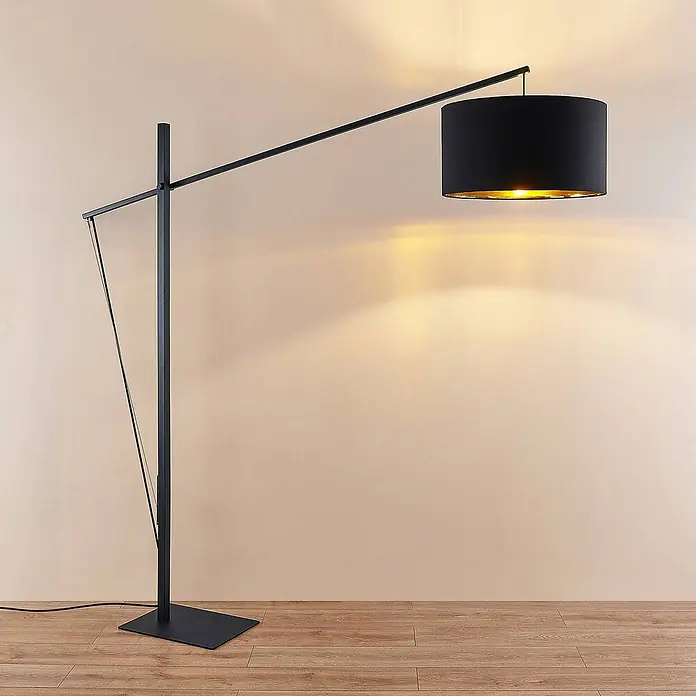 Arcchio Stehleuchte Mossa - schwarz, schwarz, E27 - Moderne schwarze Bogenstehlampe mit zylindrischem schwarzem Schirm und goldenem Inneren, die warmes Licht auf eine helle Wand wirft.